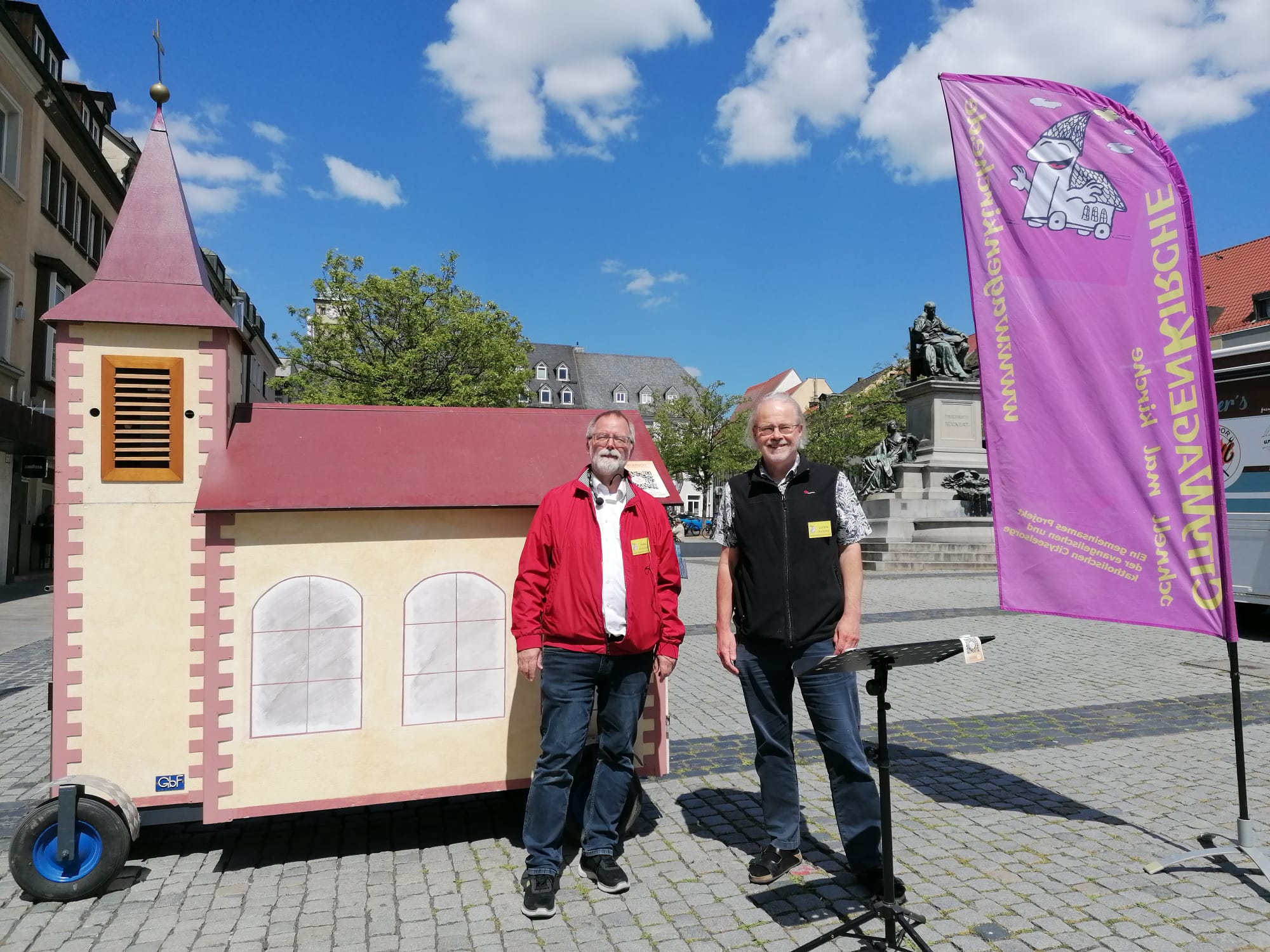 Die Wagenkirche, eine fahrbare Kirche aus Holz. Links der Turm. Der Dachfirst des "Kirchenschiffs" ist etwa so hoch wie die zwei Männer, die davor stehen: Heiko Kuschel und Lorenz Hummel. Das Foto ist auf dem Schweinfurter Marktplatz aufgenommen. Durch einen Baum sieht man im Hintergrund den Turm von St. Johannis. Rechts das Rückert-Denkmal und eine Beachflag der Wagenkirche