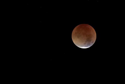 Man sieht einen schwach rot leuchtenden Vollmond am dunlen Nachthimmel, der fast vollstaändig im Erdschatten verschwunden ist.